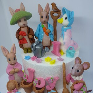 May include: A white cake decorated with a Peter Rabbit theme. The cake features figurines of Peter Rabbit, Benjamin Bunny, Jemima Puddle-Duck, and a mouse. The cake is decorated with flowers, a watering can, a rake, and a broom. The cake is also decorated with the name "Joy".
