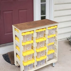 May include: A light-coloured wooden storage unit on wheels featuring three vertical columns, each holding four clear plastic bins with bright yellow lids. The unit has a flat, dark-stained wooden top surface and a sturdy, open-frame construction.