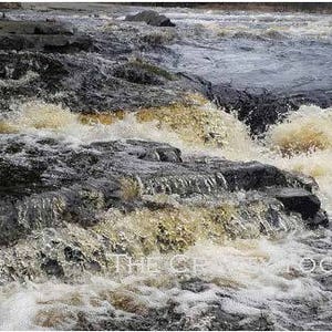 May include: A close-up of a rushing river with white water rapids flowing over dark gray rocks. The water is clear and the rocks are wet.