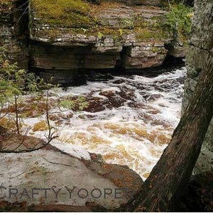 May include: A close-up view of a rushing river flowing through a rocky canyon. The water is a light brown color and is churning and bubbling. The rocks are covered in moss and lichen. The image is taken from a low angle, looking up at the river.