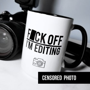 May include: White coffee mug with black text that reads "F*ck Off I'm Editing" with a camera icon below the text. The mug has a black handle and a black rim. The mug is sitting on a white surface in front of a black camera with a flash.