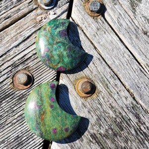 May include: Two green and pink crescent moon shaped stones with a speckled pattern. The stones are on a weathered wooden surface.