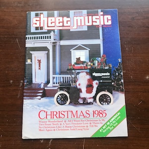 May include: A vintage-style car with Santa Claus driving, decorated with Christmas ornaments and a Christmas tree. The car is in front of a house with a red door and a snowy yard. The cover of a sheet music magazine titled "Sheet Music" with the text "Christmas 1985" and "#1 Hit in America! Saving All My Love For You".