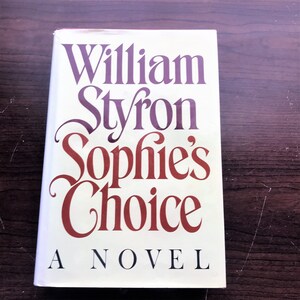 May include: A white book cover with the title "Sophie's Choice" in red and purple lettering. The author's name, "William Styron", is printed in purple lettering above the title. The words "A Novel" are printed in black lettering below the title.