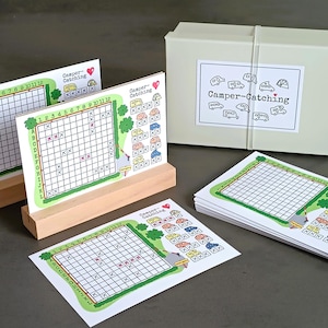 Könnte beinhalten: Ein Set von Camper-Catching-Spielkarten mit einem Raster, Zahlen und Buchstaben, präsentiert auf Holzständern und einem Stapel. Die Karten haben einen grünen Rand mit Wohnwagen-Illustrationen und dem Titel des Spiels. Eine Box mit dem Titel des Spiels ist ebenfalls enthalten.