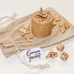 Könnte beinhalten: Ein Holzspielset bestehend aus einem zylindrischen Behälter mit nummerierten Löchern, kleinen Holzstiften, zwei sechsseitigen Würfeln und einer wohnwagenförmigen Holzfigur. Ein weißes Etikett am beigen Stoffbeutel trägt den Text Camper gewinnt!