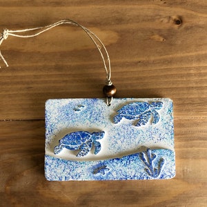 Könnte beinhalten: Ein weißer Keramik-Anhänger mit einem blauen und weißen Sprühlack-Design. Zwei Meeresschildkröten sind auf den Anhänger geprägt, eine über der anderen. Der Anhänger hat einen braunen Bindfaden-Aufhänger.
