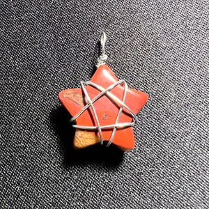May include: A red gemstone star pendant with a silver wire wrapped around it. The star is a five-pointed star with a wire design that creates a six-pointed star within it.