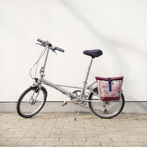 Könnte beinhalten: Ein silbernes Klappfahrrad mit schwarzem Sattel und Lenker. Es hat eine Tasche mit Blumenmuster am Heck. Das Fahrrad steht auf einer Ziegeloberfläche vor einer weißen Wand.