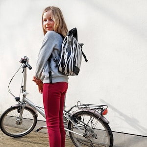 Op de afbeelding: Een persoon draagt een zwart-witte zebra-gestreepte rugzak met een zwarte basis. De persoon draagt een grijze trui en een bordeauxrode broek. Een zilveren vouwfiets staat naast de persoon.