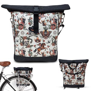 May include: A roll-top bicycle bag with a white background and a repeating pattern of cowgirls, skulls, cacti, and horses. The bag has a black top and bottom, a black buckle, and a black strap. The bag is shown attached to a bicycle.