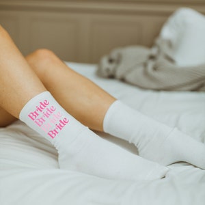 May include: White socks with the word "Bride" repeated four times in pink lettering. The socks are on a white bed.