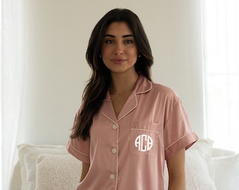 May include: A pink satin pajama set with short sleeves and shorts. The top has a white trim and a button-up front. The shorts have a white trim and a pocket on the left side with a white monogram "ACB".