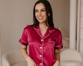 May include: A woman wearing a pink satin pajama set with white piping. The set features a short-sleeved top with a button-up front and a matching pair of shorts.