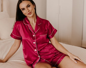 May include: A woman wearing a pink satin pajama set with white piping. The set features a collared shirt with short sleeves and matching shorts.