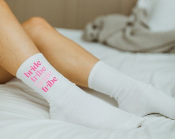 May include: White socks with the words "bride tribe" written in pink on them. The socks are on a white bed.