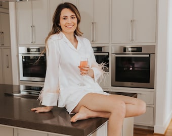 May include: A woman in a white satin pajama set with feather trim sits on a kitchen island, holding a glass of pink drink. The kitchen has white cabinets and a wood floor.