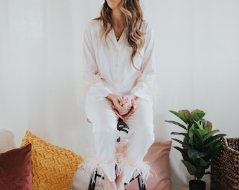 May include: A light pink pajama set with feather trim on the sleeves and pant cuffs. The pajamas are button-down and are worn by a person sitting on a black metal stool. Decorative pillows and a plant are in the background.