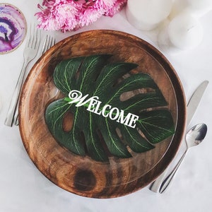 May include: A wooden plate with a green leaf with the word "WELCOME" on it. The plate is set on a white tablecloth with pink flowers and white candles.
