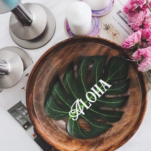 May include: A wooden bowl with a green leaf design and the word "ALOHA" written on it. The bowl is on a white surface with candles and coasters.