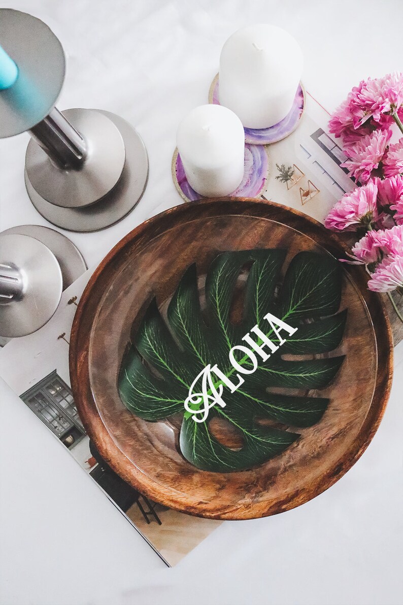 May include: A wooden bowl with a green leaf design and the word "ALOHA" written on it. The bowl is on a white surface with candles and coasters.