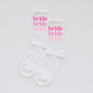 May include: A pair of white socks with the word "bride" printed on them in pink letters. The word "bride" is repeated three times on each sock.