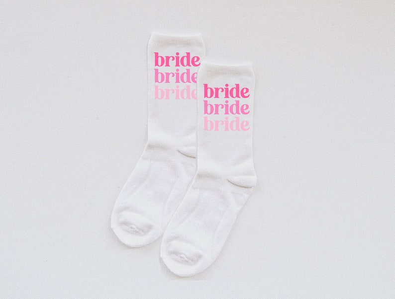 May include: A pair of white socks with the word "bride" printed on them in pink letters. The word "bride" is repeated three times on each sock.