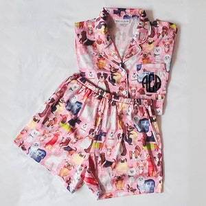 May include: Pink pyjama set with a short-sleeved button-up top and matching shorts. Both pieces feature a print of various dog breeds. The top has a monogrammed patch on the left chest that reads "ACD".