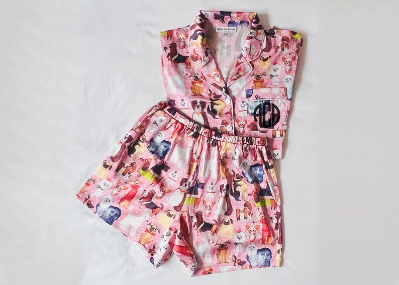 May include: Pink pyjama set with a short-sleeved button-up top and matching shorts. Both pieces feature a print of various dog breeds. The top has a monogrammed patch on the left chest that reads "ACD".