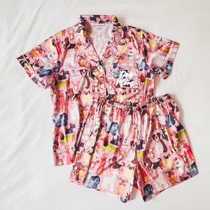 May include: Pink pyjama set with a short-sleeved top and shorts. The top has a button-up collar and a pocket with the text "Dog Mom" on it. Both the top and shorts have a print of various dog breeds.