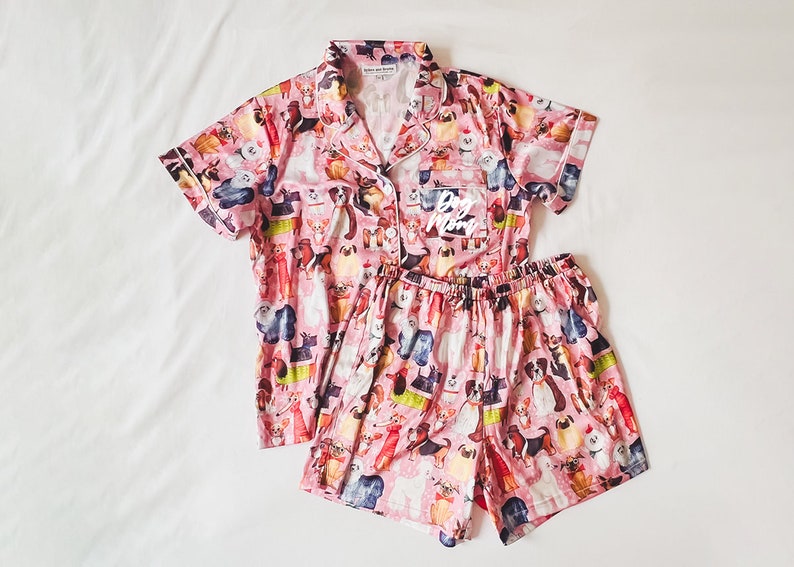 May include: Pink pyjama set with a short-sleeved top and shorts. The top has a button-up collar and a pocket with the text "Dog Mom" on it. Both the top and shorts have a print of various dog breeds.