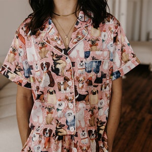May include: Pink satin pyjamas with a short-sleeved top and a dog print pattern. The pyjamas have white piping around the collar and sleeves.