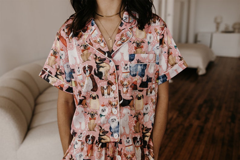 May include: Pink satin pyjamas with a short-sleeved top and a dog print pattern. The pyjamas have white piping around the collar and sleeves.