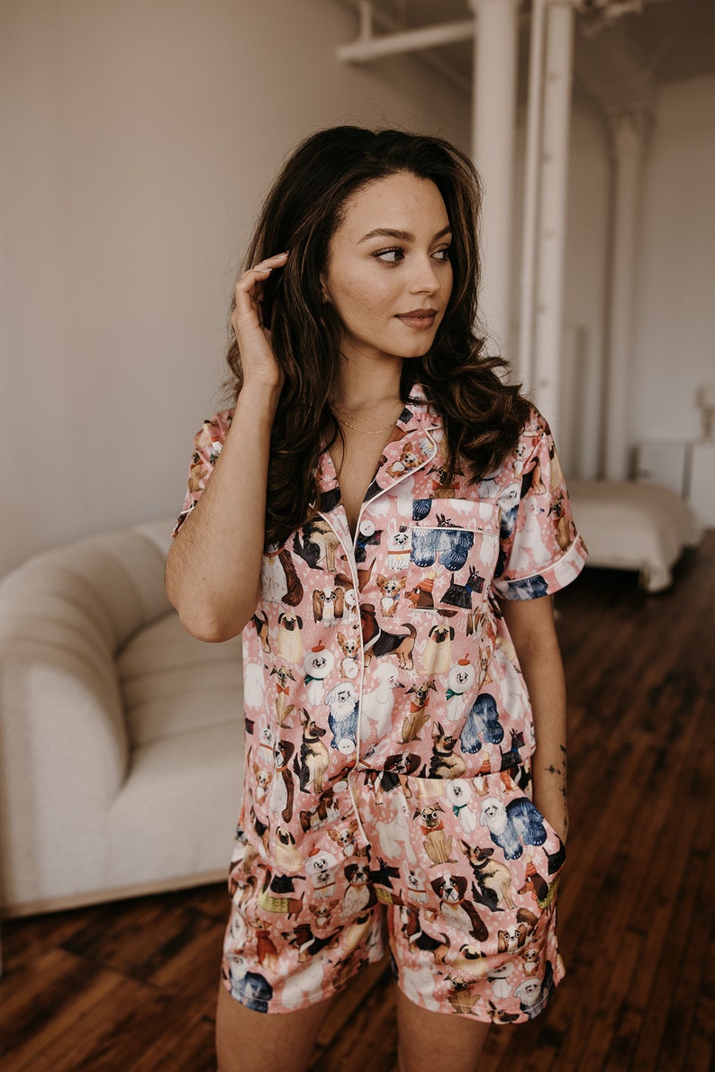 May include: A pink satin pyjama set with a short-sleeved top and shorts. The fabric features a print of various dog breeds in different poses.