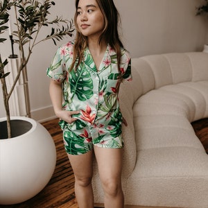 May include: A two-piece pajama set featuring a short-sleeved button-down shirt and matching shorts. The fabric is light green with a vibrant tropical print of large dark green monstera leaves and bright pink hibiscus flowers.