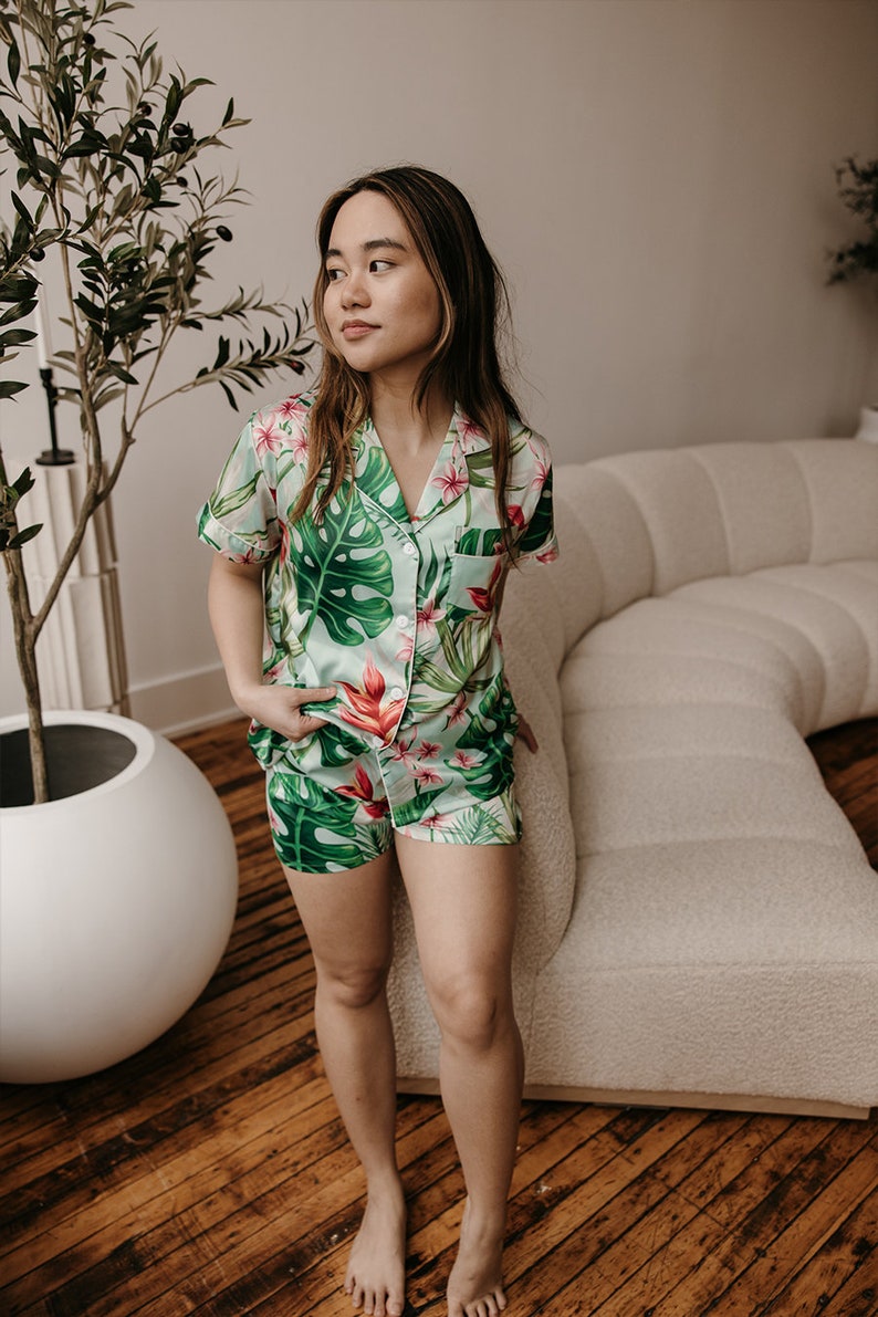 May include: A two-piece pajama set featuring a short-sleeved button-down shirt and matching shorts. The fabric is light green with a vibrant tropical print of large dark green monstera leaves and bright pink hibiscus flowers.