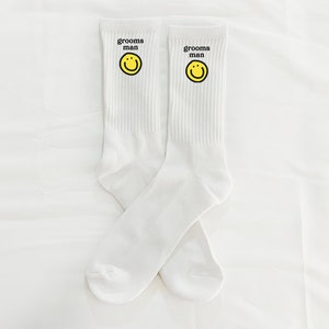 Puede incluir: Un par de calcetines blancos con una carita sonriente amarilla y el texto "grooms man" en cada calcet&iacute;n.