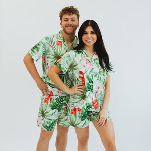 May include: Two matching pajama sets featuring a light green base with a vibrant tropical print of large dark green monstera leaves and bright red hibiscus flowers. Each set includes a short-sleeved button-down shirt with a notched collar and matching shorts.