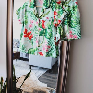 May include: A two-piece pajama set featuring a short-sleeved button-down shirt with a collar and a chest pocket, paired with matching shorts. The fabric is light green with a vibrant tropical print of large monstera leaves and red flowers.