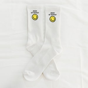 Puede incluir: Un par de calcetines blancos con una carita sonriente amarilla y el texto "man of honor" en cada calcet&iacute;n.