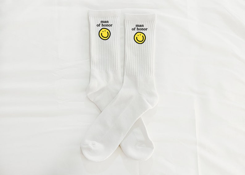Puede incluir: Un par de calcetines blancos con una carita sonriente amarilla y el texto "man of honor" en cada calcet&iacute;n.
