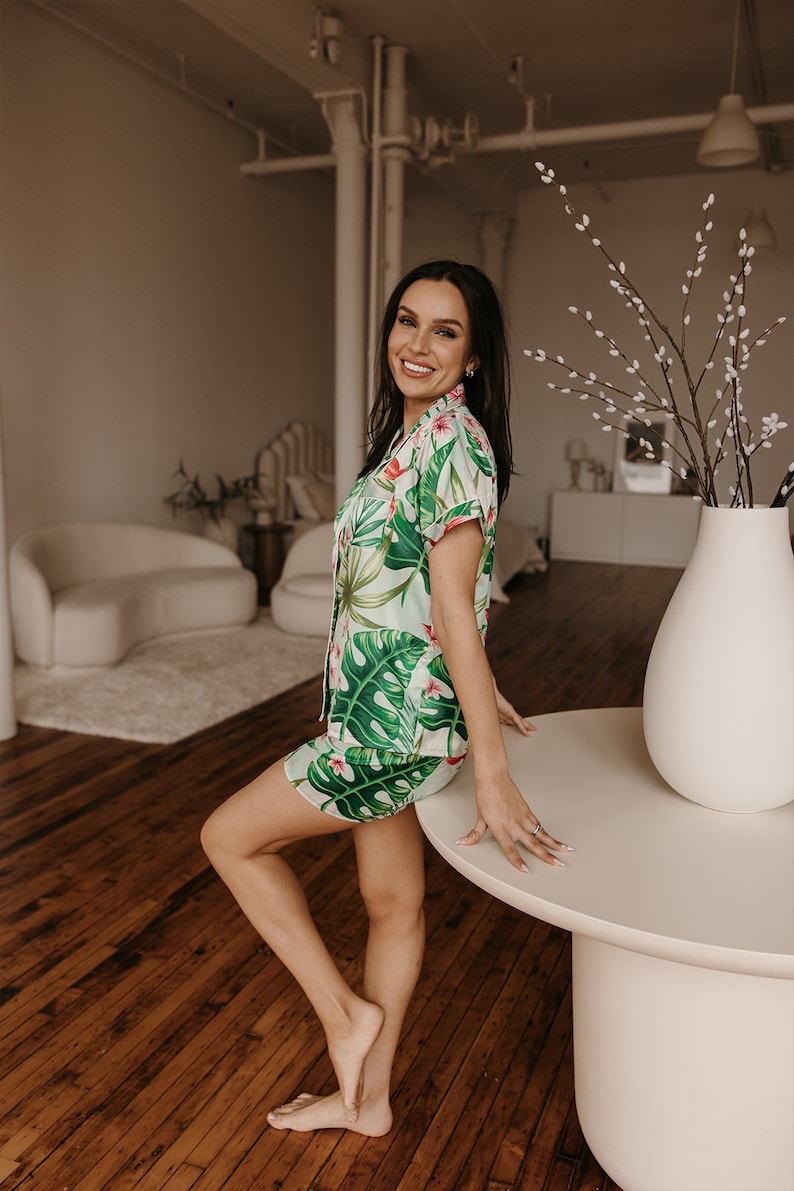 May include: A two-piece pajama set featuring a short-sleeved button-down shirt and matching shorts, decorated with a vibrant green tropical monstera leaf and pink floral pattern on a light cream-colored fabric with pink piping trim.