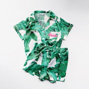 May include: White pajamas with a green tropical leaf print. The top has a button-up closure and a pocket with the word "Bride" embroidered in pink.