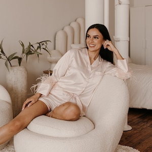 May include: Light pink satin pajama set with feather trim on the sleeves and shorts. The set includes a button-down top and shorts. The model is sitting in a cream-colored chair.