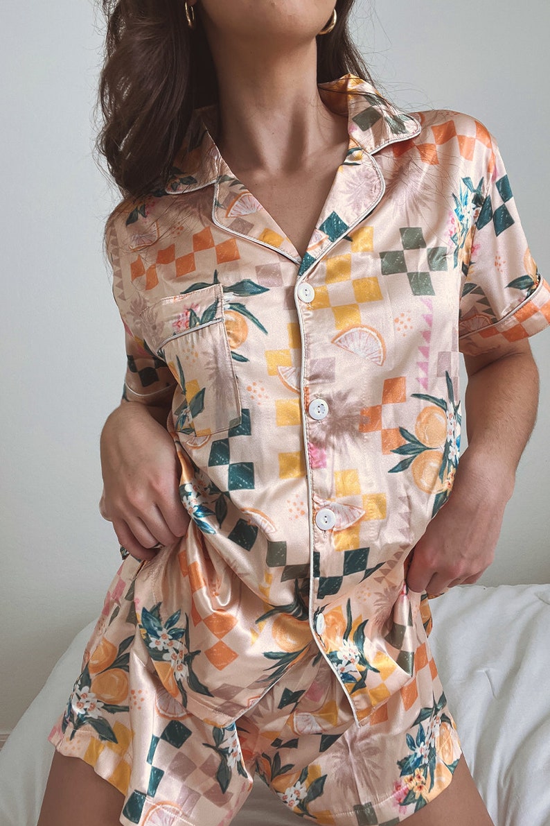 May include: A peach-coloured satin pyjama set with a checkered pattern and a citrus fruit print. The set includes a short-sleeved button-up top and matching shorts.