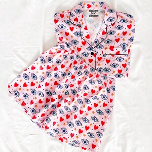 Puede incluir: Conjunto de pijama rosa con una parte superior de manga corta abotonada y pantalones cortos. La tela presenta un patrón repetido de ojos azules, corazones rojos y estrellas doradas. La parte superior tiene un cuello y el texto "Splices and Seams" impreso en la parte delantera.