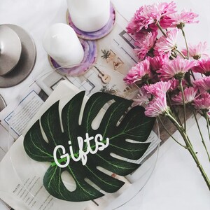 May include: A clear glass plate with a green monstera leaf cutout on top. The leaf has the word "Gifts" written on it in white. Pink flowers and white candles are in the background.