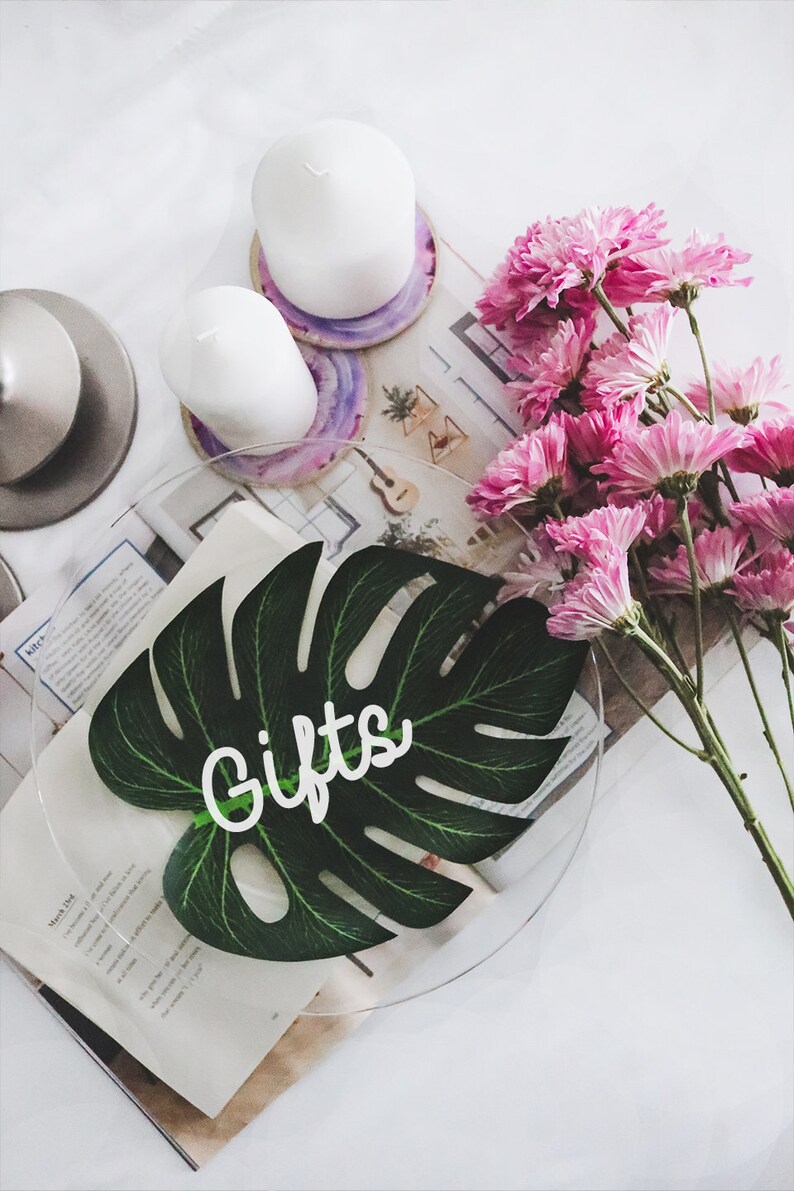 May include: A clear glass plate with a green monstera leaf cutout on top. The leaf has the word "Gifts" written on it in white. Pink flowers and white candles are in the background.