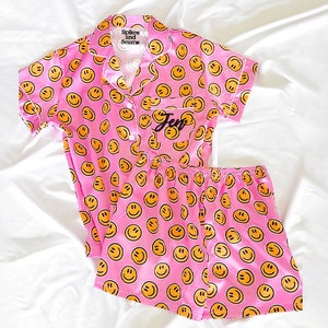 以下が含まれることがあります： ピンクのサテンパジャマセット。半袖トップとショートパンツ。トップはボタンダウンカラーで、ポケットには「Spikes and Sprouts」の文字が書かれています。セットは黄色のスマイリーフェイス柄で覆われています。ポケットには「Jen」の文字が刺繍されています。