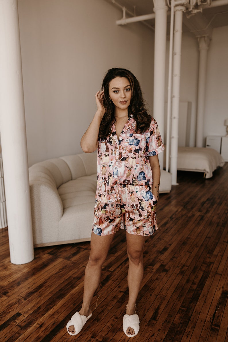 May include: A woman wearing a pink and blue short-sleeved pyjama set with a dog print. The pyjamas have a button-up front and a relaxed fit. The woman is standing on a wooden floor and is wearing white fluffy slippers.